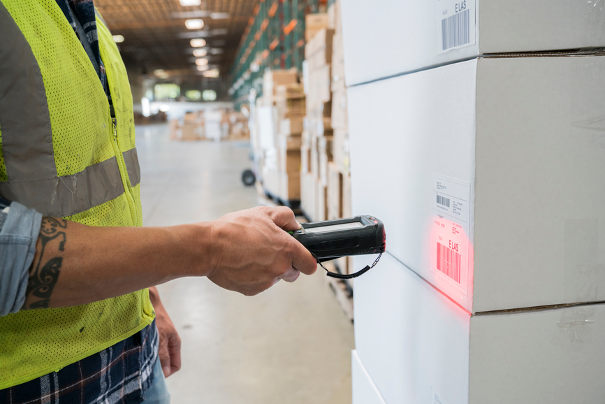 Warehouse worker  wearing a reflective vest and using a hand held RF scanner to check shipping lable bar codes on cardboard boxes full of inventory.